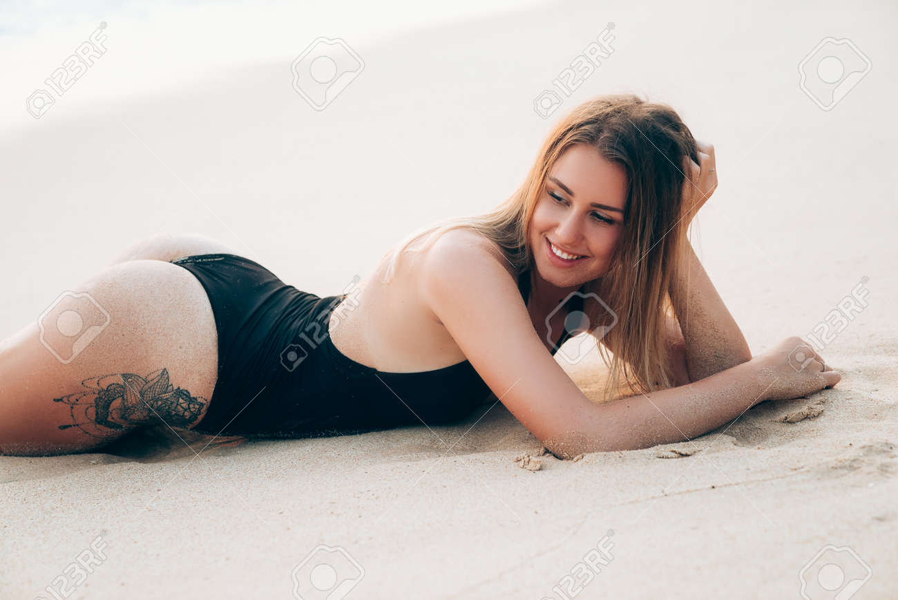 Close-up of a young beautiful girl lies on white sand in a black swimsuit, propps her head with her hand, counts with her friends, enjoys sunburn, has sexy tatu.Concepuia active rest. - 105288500