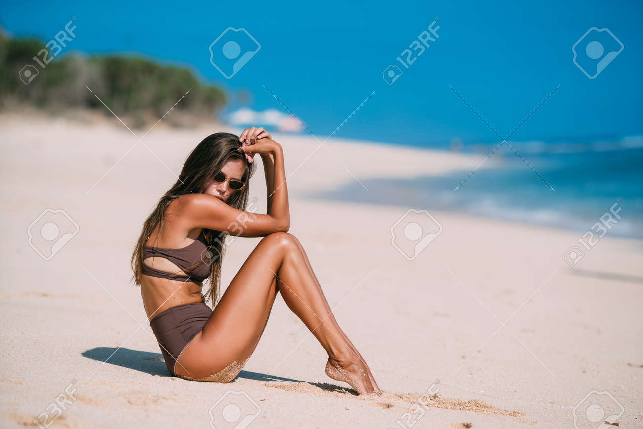 Sexy model in brown swimsuit and sunglasses posing on white sandy beach - 119905036