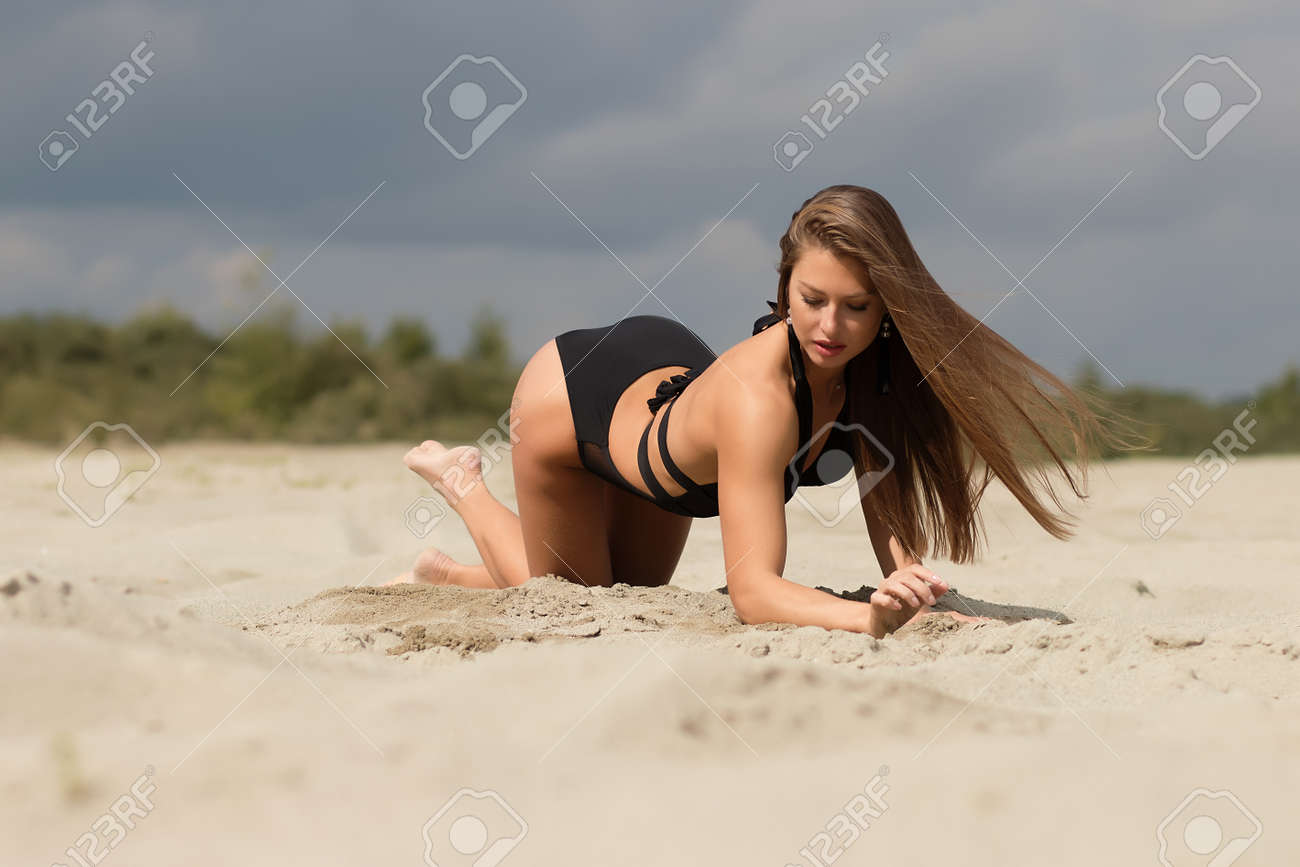 Seductive slim woman in trendy black swimsuit and earrings standing on all fours on sand under cloudy sky looking at camera - 142619464