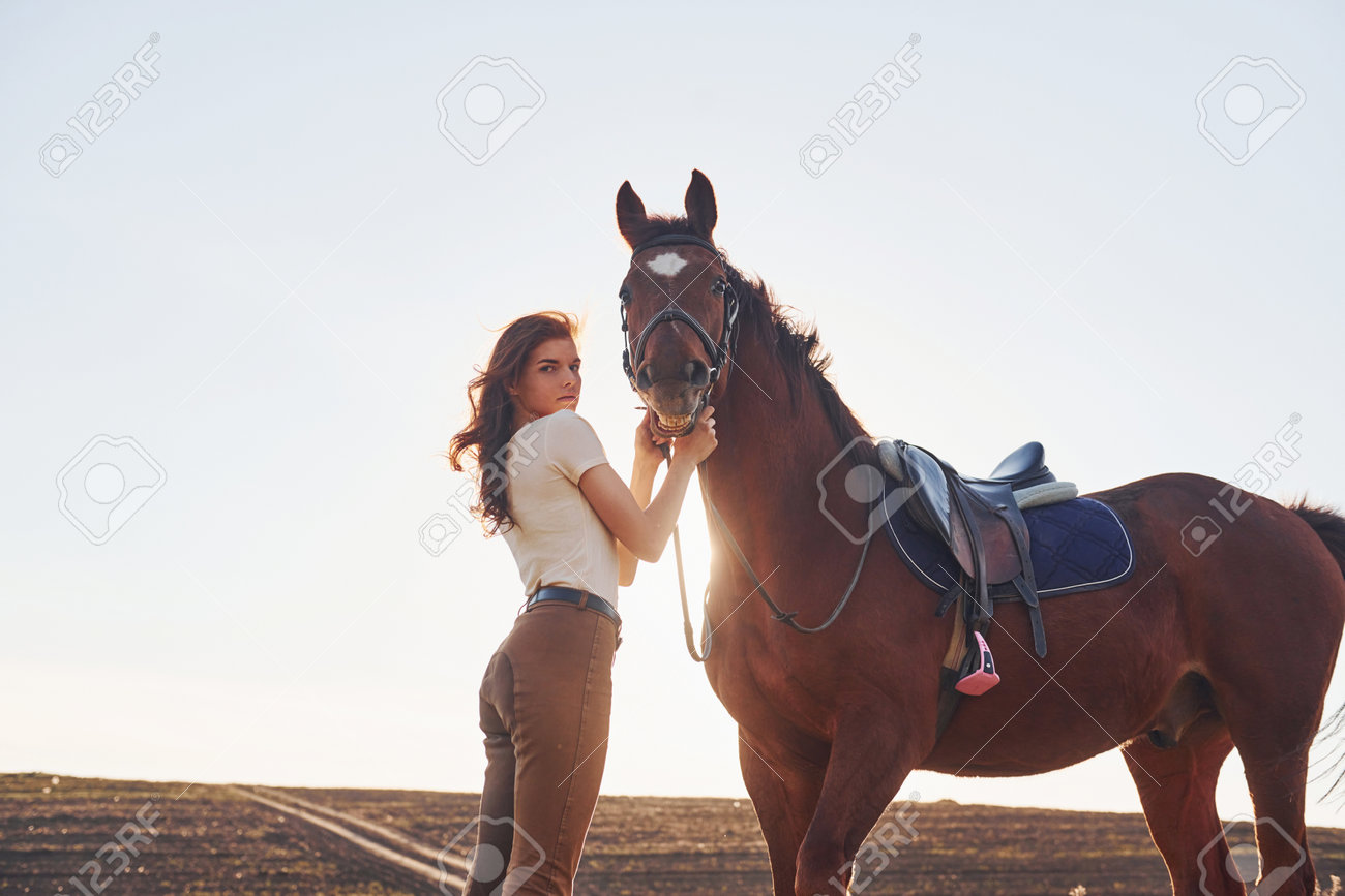 Beautiful sunshine. Young woman standing with her horse in agriculture field at daytime - 201419048